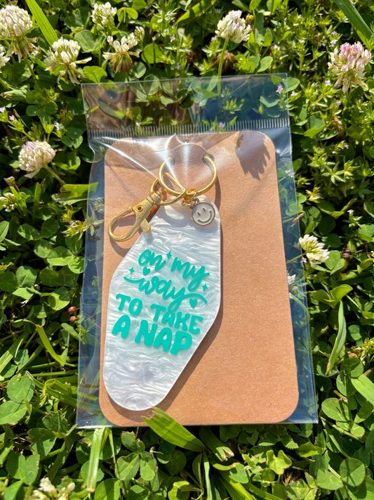 handmade motel-style acrylic keychain with pearlescent white tag, teal lettering that reads on my way to take a nap, gold clasp and smiley face charm