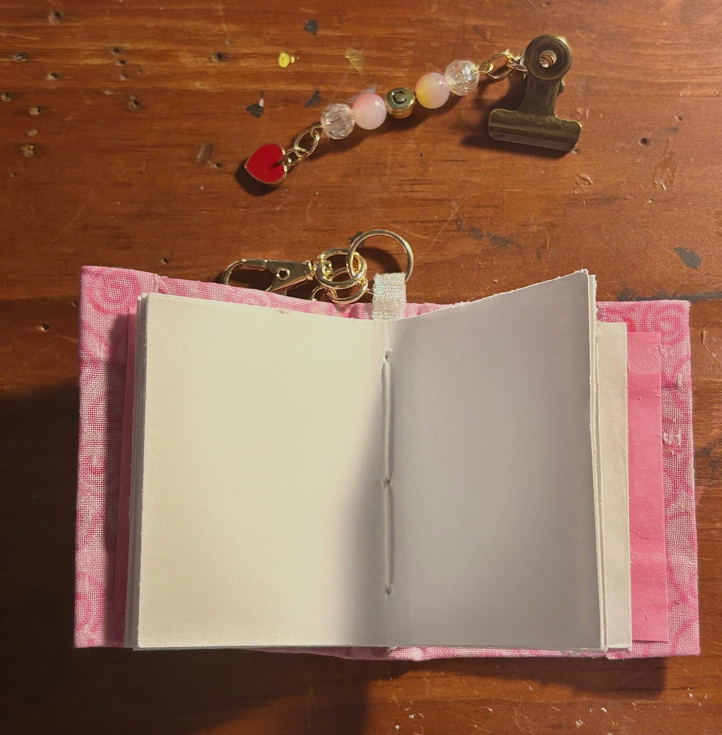 handmade mini journal open to blank deckle-edge pages, pink floral fabric cover, with a beaded keychain charm and initial binder clip charm